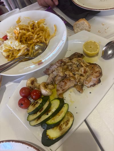 The image shows a close-up of a meal served on white plates, set on a white tablecloth. In the foreground, a white rectangular plate contains a grilled pork chop topped with sliced garlic and what appears to be mushrooms. To the left of the pork chop are cherry tomatoes and sautéed mushrooms. Below the pork chop and vegetables, grilled zucchini slices are arranged. A half-lemon is also visible on the plate. To the left and slightly behind this plate, another white bowl contains a serving of french fries mixed with what looks like scrambled eggs and red bell peppers. A spoon is resting in this bowl. In the background, a slice of bread sits on a folded white napkin. A portion of a person's hand is visible in the top center of the image. The lighting suggests an indoor setting, possibly a restaurant. The overall scene depicts a prepared meal ready to be eaten. The specific location of Urbanización Roquetas De Mar, Spain is not evident from the visual information provided.