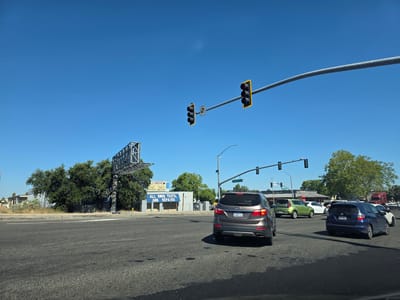 The image depicts a sunny daytime scene at a busy intersection in West Sacramento, California. The sky is a clear, bright blue, suggesting fair weather. The foreground is dominated by an asphalt road, marked with white lines, indicating multiple lanes of traffic.
Several vehicles are present, including a gray SUV in the right lane, a blue hatchback behind it, and a lime green car in the left lane, all seemingly stopped at a red light. Further down the road, other cars and a red semi-truck are visible, suggesting ongoing traffic flow.
Above the intersection, traffic lights hang from a large metal pole that spans across the road. One set of traffic lights shows red for all bulbs, while another set, further to the left, also shows red. A street sign indicates "Carmichael Ave."
To the left of the intersection, a large industrial-style billboard structure stands tall. Adjacent to it is a building with a blue banner that reads: "ALL SMOG TESTS, AND REPAIRS." The building appears to be a business, possibly a smog testing and repair shop. The overall scene conveys the everyday hustle and bustle of a suburban or commercial area. There are no people visible in the image.