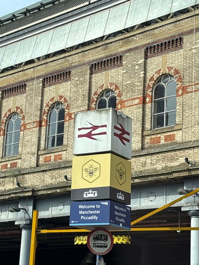The image shows a vertically oriented view of a signpost at Manchester Piccadilly train station in Manchester, United Kingdom. The signpost is comprised of stacked cubes with the National Rail logo on the top cubes, a bee symbol (a symbol of Manchester) on the next cube down, and "Welcome to Manchester Piccadilly" on the bottom cube. Behind the sign is the station's brick exterior with arched windows. A weight limit sign is also visible next to the signpost. The image is rotated.