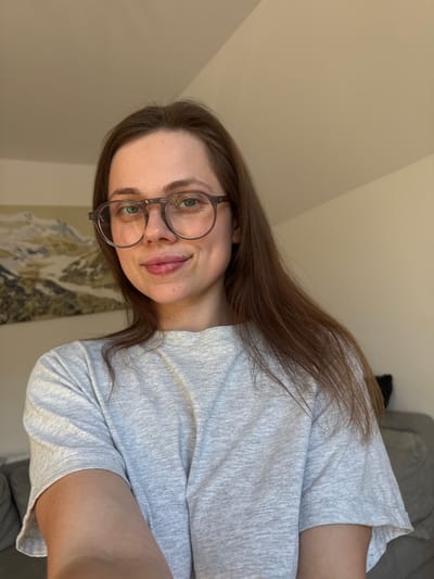 This is a selfie of a young woman with long, brown hair and glasses. She is wearing a grey t-shirt. Her face is turned to the side, and she is smiling. The background is a plain, light-colored wall, with a blurry image of what appears to be a landscape painting or photograph hanging on it. The lighting suggests it might be daytime, with natural light coming from the side. The orientation of the photo is vertical, and the woman is positioned in the right half of the frame.