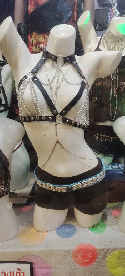 This image shows a display in a shop in the Ko Pha-ngan District of Thailand, likely a clothing store or boutique selling festival wear. The primary subject is a white mannequin dressed in a black harness-style top adorned with multiple silver chains. The harness is connected to black shorts that feature a decorative belt made of small white shells and blue beads. A delicate silver body chain is draped over the mannequin's torso. In the background, other mannequins are visible, some wearing jewelry and other clothing items. The display is set against a backdrop that includes patterned fabric and glass, suggesting a retail environment. The flooring beneath the display is covered with a white material featuring colorful circular polka dots in shades of yellow, blue, pink, and red. The overall impression is of a vibrant and eclectic collection of fashion items.