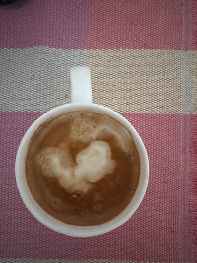 A top-down, eye-level shot shows a white coffee mug filled with a frothy, brown beverage, possibly coffee or hot chocolate, resting on a striped textile surface. The textile features bands of pink, white, and light blue. The beverage in the mug has a heart-shaped pattern created by the foam. The scene is static, with no people or discernible action. The overall lighting is soft and even. There is no information available to determine the location in the city of Glasgow, United Kingdom.