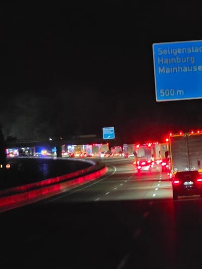 The image depicts a nighttime scene on a German highway, specifically near Seligenstadt. A blue highway sign indicates "Seligenstadt," "Hainburg," and "Mainhausen" with "500 m" below, signifying the distance to these locations. The highway is congested with traffic, including multiple emergency vehicles with flashing red and blue lights, suggesting an incident or accident. Several trucks and cars are visible, their headlights and taillights illuminated. The road surface is wet, reflecting the lights and the overhead structures.