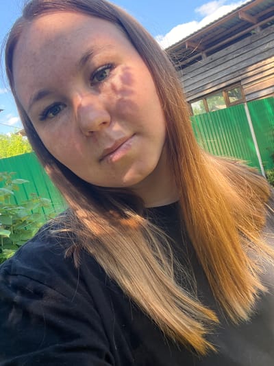 A young woman with long, auburn hair, wearing a black t-shirt, is taking a selfie outdoors. She has green eyes and light skin with freckles. Her hair is styled in loose waves and extends down her shoulders. The background features a rustic wooden building, a vibrant green corrugated metal fence, and lush green foliage. The sky above is clear blue with scattered white clouds. Sunlight casts shadows on her face, indicating it is daytime. The overall setting suggests a rural or suburban environment, possibly in the city of Aryazh, Russia, given the context provided.