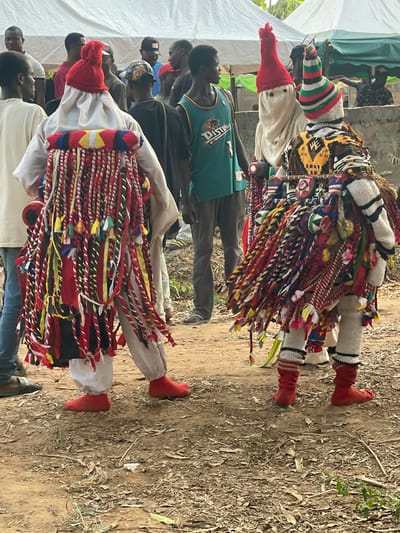 The image displays an outdoor scene in Umueze, Nigeria, featuring two elaborately costumed figures observed by multiple unmasked individuals.
The figure on the left is predominantly dressed in white, comprising a tunic and trousers, accented by a solid red conical hat and solid red foot coverings. A rigid frame on its back is extensively covered with long, braided strips of fabric in red, white, yellow, and black, extending downwards. A red, cylindrical object is partially visible on its left hip.
The figure on the right wears a multi-patterned tunic in yellow, black, and white, with white knitted sleeves and leggings. Its head is covered by a conical hat featuring horizontal stripes of red, white, green, and black. Its feet are covered in red material extending above the ankles, distinct from the left figure's foot coverings. A similar frame on its back is densely adorned with braided fabric strips and woven textiles in red, blue, green, yellow, and white, some ending in small tassels.
Both masked figures stand on uneven, dry earthen ground, sparsely covered with leaves and small plant debris. In the background, approximately ten to fifteen unmasked adult males are present, observing the scene. One man in the mid-ground wears a teal basketball jersey with the word "DISTONS" and the number "7." Partially visible in the upper background are elements of white canopy tents and some distant foliage, indicating an outdoor gathering. The scene is illuminated by natural daylight.