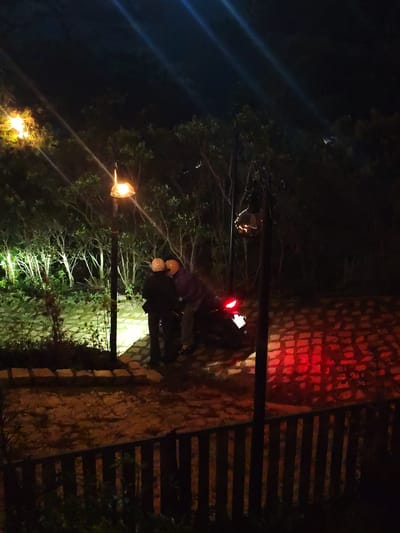 This nighttime photograph, taken in Lạc Dương, Vietnam, captures two individuals standing beside a motorcycle on a pathway surrounded by trees.
The primary subjects are two people, both wearing helmets – one cream/white and the other yellow/orange. One individual, dressed in dark attire, stands upright near a lamppost on the left. The second person, wearing a purple top and lighter-colored pants, is leaning towards a dark-colored motorcycle. The motorcycle's red taillight is illuminated, casting a glow onto the textured brick ground behind it.
The scene is set outdoors, characterized by dense vegetation and trees that frame the pathway. The ground surface varies, with areas of dirt or gravel and sections of patterned brick or stone paving. Two streetlights provide illumination: one on the left brightly lights the area around the people and some trees, while another on the right features a more decorative fixture. Bright lens flares from these light sources are visible against the dark sky, emphasizing the nighttime setting. A dark wooden fence runs across the foreground, indicating a boundary.
The activity appears to be a quiet interaction between the two individuals, possibly a conversation or a moment of preparation or pause with the motorcycle. The presence of helmets suggests they are motorcyclists. The time of day is clearly night, evidenced by the darkness and reliance on artificial lighting. No discernible text is visible in the image. The mood is calm and intimate, highlighted by the focused light on the subjects amidst the dark surroundings.