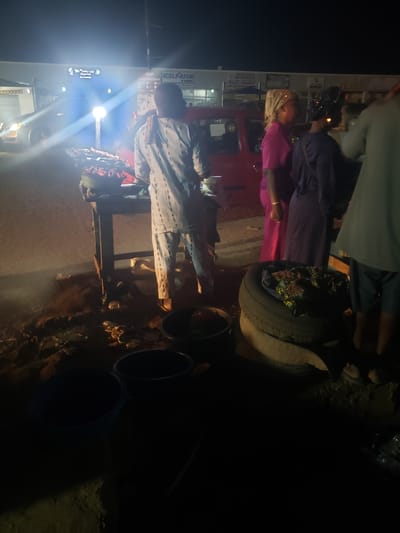 At night in Ibadan, Nigeria, a group of people are gathered around a street vendor. The vendor is positioned in front of a shop with signage for “ICELIQUOR” and “The Success Space”. A bright light shines from a vehicle near the vendor. A red vehicle is near the group. Some people are dressed in traditional African clothing, with one person wearing a pink outfit and gold headwrap. The vendor's setup includes black buckets, a wooden table, and what appears to be a tire being used as a surface. The ground is littered with debris.