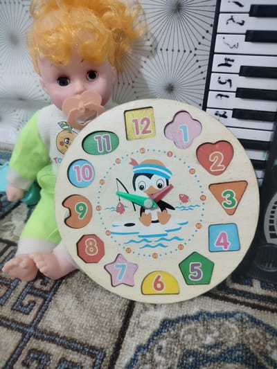 A doll with blonde hair and a pacifier is sitting on a patterned rug. In front of the doll is a wooden clock puzzle with numbers and shapes. To the right of the doll is a keyboard with black and white keys. The background consists of a white surface with a geometric pattern. The scene is indoors, likely in a domestic setting, but no specific city or country can be identified from the visual information provided. The doll is positioned with its legs extended, and the clock puzzle is tilted towards the viewer.