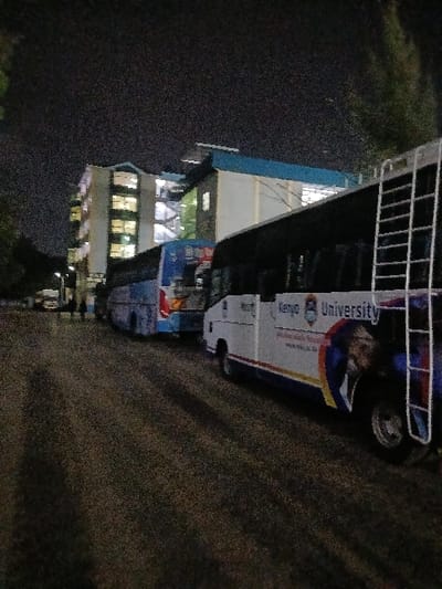 The image depicts an outdoor nighttime scene within Thika Township ward, Kenya, specifically near Kenyatta University facilities. A dark, textured road forms the foreground. On the right, a white bus bearing "Kenyatta University" branding and a visible logo is parked, equipped with a white ladder mounted on its rear-right side. Partially obscured behind it to the left is a blue bus. In the background, a multi-story, light-colored building stands illuminated by numerous interior lights visible through its windows, featuring a prominent blue roof or architectural accent. A smaller, adjacent building with similar blue detailing is partially visible to the right of the main structure. A few indistinct human figures are observed near the buildings in the far distance. The sky is dark, consistent with nighttime, and artificial lights from the buildings and distant streetlights illuminate the environment. Sparse foliage is present along the scene's edges.