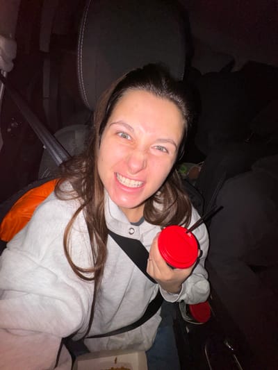 A woman grins at the camera, her teeth bared. She wears a light gray hooded sweatshirt and a seatbelt. She holds a red-lidded cup. A second red-lidded cup is in the background. She appears to be in a car, given the seatbelt and surrounding upholstery. The lighting is dim.
