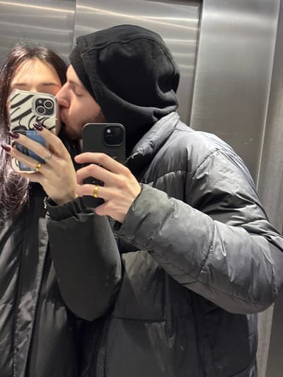 The image is a mirror selfie featuring two individuals, rotated clockwise from a typical upright orientation. The subjects are captured in a close, affectionate pose, appearing to kiss or share an intimate moment in their reflections.
The individual in the upper portion of the frame wears a dark grey or black puffer jacket over a black hooded sweatshirt, with the hood pulled up. Their left hand is visible, holding a black smartphone with a distinct camera module (resembling an iPhone) to capture the selfie. A simple gold band ring adorns their ring finger. Their face, partially obscured by the phone and the reflection of the other person, shows some facial hair.
The second individual is primarily seen as a reflection in the lower part of the frame. They have long, dark hair, and their face is turned towards the first person's reflection, with their lips seemingly touching or very close. Their right hand is visible, holding a light-colored smartphone (also resembling an iPhone) encased in a clear cover with a black-and-white zebra print design visible behind it. This individual wears a silver ring on their ring finger, and their fingernails are painted with a dark red or maroon polish.
Both individuals are dressed in casual, dark-colored outerwear, suggesting a cool environment or casual setting. The background consists of a plain, light grey, highly reflective surface, likely brushed metal or a polished wall, which serves as the mirror for the selfie. The overall scene conveys a sense of intimacy and connection between the two subjects as they capture a shared moment. No visible text is present in the image.