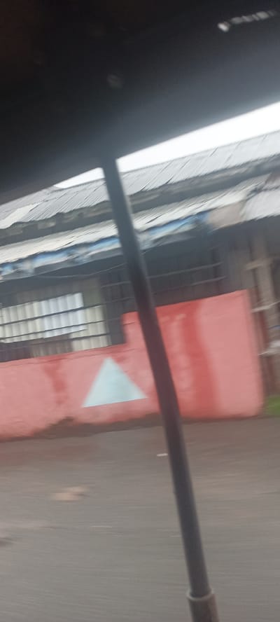 The image shows a blurry view from inside a vehicle, possibly a bus, in Umuahia, Nigeria. A portion of the vehicle's interior, a pole, is visible in the foreground. Outside, there is a building with a corrugated metal roof and a wall painted a reddish-pink color. A light blue triangle is painted on the wall. The wall appears to run along the roadside. There are bars on the windows of the building. The road surface is asphalt. The overall scene is somewhat indistinct due to the motion blur.
