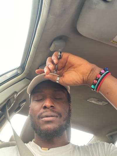 A man is sitting inside a vehicle in Galadimawa Village, Nigeria, with his eyes closed and his hand resting on his forehead. He is wearing a white t-shirt, a light-colored baseball cap, and several rings on his fingers, including two silver bands on his right hand. On his left wrist, he wears multiple bracelets, one with colorful dice-shaped beads. The interior of the vehicle is visible, including the car ceiling and a sun visor. The man has a beard and a necklace with a wooden pendant. The overall lighting suggests it is daytime.