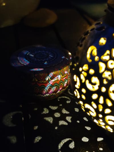 The image depicts a dimly lit indoor scene, possibly a room or an alcove. In the foreground, on the left, is a small, round, dark-colored tin with intricate colorful patterns. The tin has a geometric design with red, green, blue, pink, and yellow accents, and it reflects light. To the right of the tin is a decorative lantern with cut-out designs. The lantern is illuminated from within, casting a warm, yellow glow through its perforations onto a dark surface in front of it. This surface is also decorated with lighter, scattered cut-out patterns. In the background, out of focus, are large, rounded, light-colored objects, possibly stones or decorative elements. The overall impression is one of ambient, decorative lighting. There is no indication of the city of Pyatigorsk, Russia, or any people in the image.
