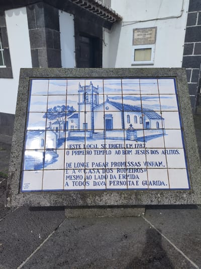 This is an outdoor scene featuring a tile mural mounted on a gray, weathered stone base. The mural depicts a church and associated buildings in a blue and white design. Below the illustration, there is text in Portuguese which reads: "NESTE LOCAL SE ERIGIU EM 1787, O PRIMEIRO TEMPLO AO BOM JESUS DOS AFLITOS. DE LONGE PAGAR PROMESSAS VINHAM, E A «CASA DOS ROMEIROS» MESMO AO LADO DA ERMIDA A TODOS DAVA PERNOITA E GUARIDA." In the bottom left corner of the mural, "E.VIEIRA, LAGOA, S.MIQUEL, Reones" is visible, suggesting the artist or location reference. The mural is framed by a weathered gray stone.
Behind the mural, there is a building with white walls and dark stone corner detailing. A sign is mounted above the mural on the building's wall; "SENAL ROMELES PRO & SHESPOSA FRANCISCA MABA ALFEDRO, 1787" can be seen. The scene appears to be in the city of Ponta Delgada, Portugal, based on the language and artistic style of the tile mural. The ground in front of the mural is paved with black stones.