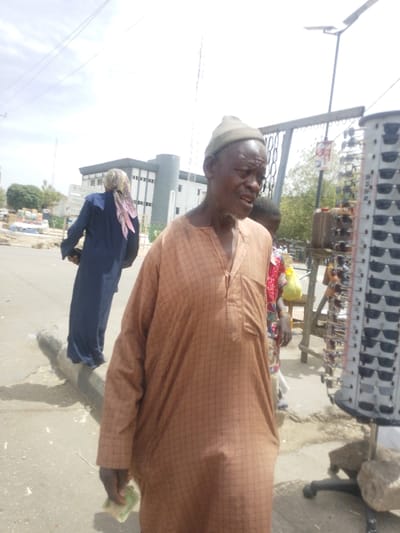 An outdoor scene in Gyadi Gyadi Arewa, Nigeria, depicts an elderly man in the foreground, positioned right of center, looking slightly leftward with his mouth slightly open. He wears a light brown, grid-patterned kaftan and a matching knitted cap, holding a folded green banknote in his right hand. To his immediate right is a tall, black-framed rotating display stand exhibiting numerous pairs of sunglasses. Behind him, a partially visible child in a red and white patterned garment stands near a makeshift stall with a brown jerry can. Further in the mid-ground on the left, a person dressed in a dark blue gown and a pink-patterned head covering walks away from the viewer along a concrete curb. The environment features an asphalt road, a grey concrete curb, and in the distant background, a modern multi-story building with light and dark grey facades. The sky is bright and overcast. A street light with a solar panel is visible in the upper right.