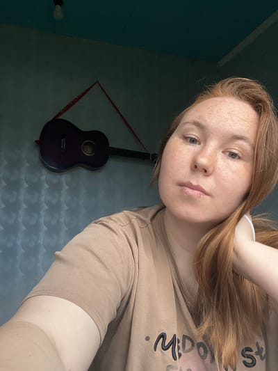 The media file features a young woman with fair skin and auburn hair, who appears to be taking a selfie. She has a light dusting of freckles across her nose and cheeks, and her eyes are a light green. She is wearing a light brown t-shirt. Her left arm is behind her head, and her hair cascades down her shoulder.
In the background, a dark-colored guitar is hanging on a wall. The wall behind the guitar is painted a muted blue and features a subtle, wave-like pattern. A red string forms a diamond shape around the guitar. To the left of the guitar, a lightbulb is visible on the blue wall.
The lighting suggests it could be daytime, possibly indoors, given the ambient light and the indoor setting. There is no specific indication of the weather or time of day beyond this. The overall mood conveyed by the image is casual and relaxed.
There is some visible text on the woman's t-shirt, partially obscured by her arm and hair. The visible letters suggest words like "Midori" and "sts" are present.
The scene appears to be a personal space, possibly a bedroom or a living area in a home in Аряж, Russia. The orientation of the image is slightly tilted.
