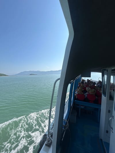 This image depicts a boat traveling on a body of water, with a clear blue sky and distant mountains visible. The perspective is from within the boat, looking outwards. On the right side of the frame, passengers are seated under a dark awning. The passengers are wearing red vests and straw hats. The water in front of the boat is churned up, indicating movement. The scene is likely a tourist excursion in or around Nam Ninh Hòa, Vietnam, given the geographical features.
