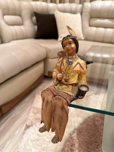A detailed, close-up, eye-level shot captures a decorative figurine of a Native American child seated on a glass surface. The figurine, made of resin, depicts a young child with dark hair adorned with a feather and a beaded headband. They wear a yellow tunic with fringe detailing and brown trousers. The child holds a dark horn-shaped object in their right hand and has a small animal figure perched on their left shoulder. In the background, a large, plush, cream-colored sectional sofa with tufted cushions and dark brown accent pillows is visible. The flooring appears to be a combination of light wood planks and a plush cream and brown rug. The scene is lit by soft, ambient lighting. There are no identifiable landmarks or contextual clues in the image that would suggest its location in Targovishte, Bulgaria.