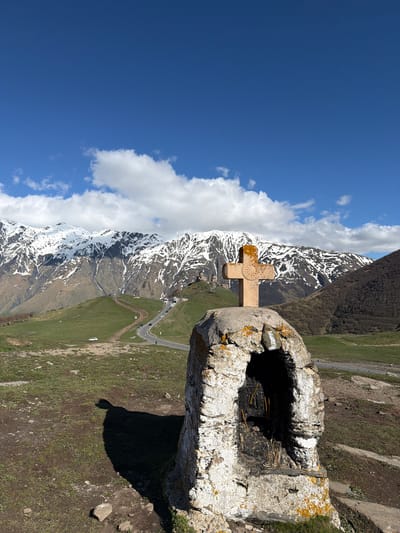 A vertical shot depicts a weathered stone cross and offering structure on a hillside in Gergeti, Georgia. The sky is bright blue with some clouds on the left. The cross is centrally positioned, superimposed against the snow-capped mountains in the background. The stone structure shows signs of age and exposure. The hillside is covered in green grass. A road snakes up the mountain, with cars on it. The lighting is bright and sunny.
