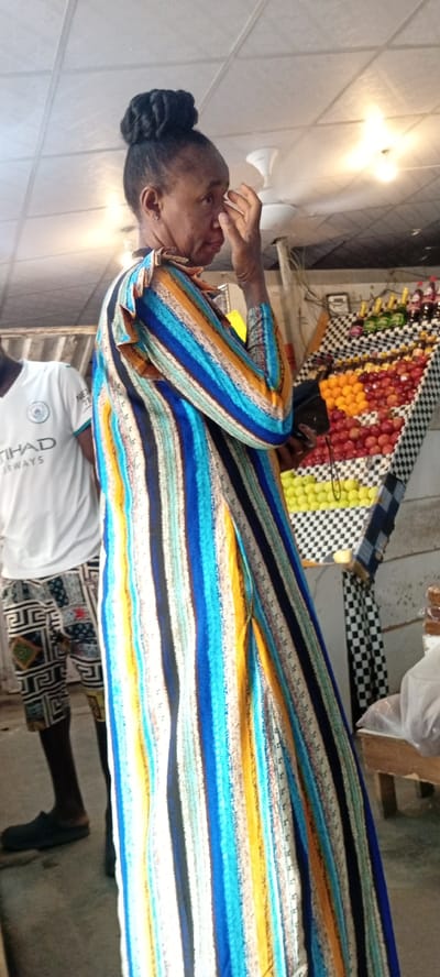 The image shows a woman standing in what appears to be a market or an outdoor shop in Aviation Quarters, Nigeria. She is wearing a long, brightly colored striped dress with blue, yellow, orange, and white stripes. Her hair is styled in a bun on top of her head. She is touching her face with her right hand, her expression is difficult to discern.

To her left, a person wearing a white t-shirt with "ETHAD AIRWAYS" written on it and patterned shorts is partially visible. In the background, a display of fruits, including oranges and apples, is arranged on shelves covered with a black and white checkered cloth. Some bottles, possibly beverages, are also visible on the top shelf. The scene appears to be outdoors, possibly during the daytime, with artificial lighting visible on the ceiling. The overall atmosphere seems to be that of a bustling marketplace.