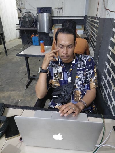 An indoor scene in Mns Alue, Indonesia, depicts an adult male seated at a light-colored table, facing the camera with a neutral expression. He is wearing a patterned short-sleeved shirt over a black t-shirt, dark trousers, a black fanny pack, a dark wristwatch on his left wrist, a silver-colored ring on his left ring finger, and multiple bracelets on his right wrist. His left index finger touches the skin below his left eye, while his right hand rests on the trackpad of an open silver Apple MacBook laptop in front of him. To the left of the laptop, a dark smartphone lies face down. To the right, another dark smartphone is connected by a white charging cable.
In the mid-ground, a second individual, wearing an orange or light brown t-shirt, is seated with their back to the camera at another dark-framed table with a light top. The background features a large dark industrial fan, a tall stainless steel machine with a digital display, and a light-colored display unit, possibly a freezer. An orange and blue plastic cup with a straw rests on the table behind the main subject. The walls are light-colored, with a horizontal black stripe running across the mid-section and a patterned area resembling black and white/grey bricks/tiles on the right. Exposed electrical wiring is visible on the walls. The floor is concrete.