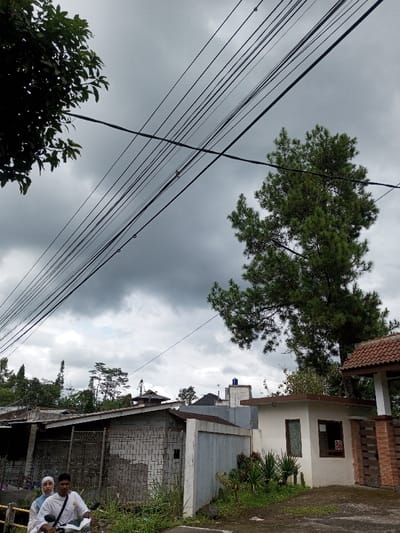 The image depicts an overcast street scene in Cibadak, Indonesia. The sky is heavily clouded and gray. Numerous black utility lines crisscross diagonally across the upper two-thirds of the frame. In the upper left, dark green foliage from a tree is partially visible, while a large, prominent conifer tree with dense green needles occupies the upper right and central right portions.

In the mid-ground, a collection of residential or commercial structures lines what appears to be a street or path. From left to right, a structure with a metal gate and unrendered blockwork is visible. Adjacent to it is a light-colored concrete wall, followed by a small, cream-colored single-story building with dark window trim and a small red sign displaying "88" next to a window. This building features a section of red-tiled roof. Further in the background, additional buildings with varying rooflines are visible, including one with a blue water tank.

In the lower-left foreground, two individuals are seated on a white scooter. The male driver, with light skin and short dark hair, wears a white long-sleeved shirt. Behind him, a female passenger wears a light-colored head covering (hijab) and matching attire. Both face towards the right. The immediate foreground consists of a dirt or gravel path with patches of green grass and foliage. A low barrier or wall is present in front of the scooter.