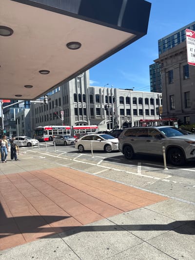The image depicts a street scene in San Francisco, California, under a clear blue sky. In the foreground, a red brick sidewalk runs diagonally across the frame, transitioning to a gray, speckled sidewalk. A large overhang with circular lights extends from an unseen building on the left, casting a shadow on the sidewalk.
The street is bustling with traffic. A red and white bus with "20" displayed prominently on its front is stopped at what appears to be a bus stop. Several cars are visible on the road, including a white sedan, a silver sedan, and a gray SUV. Pedestrians are present on the sidewalk, two women walking together.
In the background, various buildings line the street. Some have an older, ornate architectural style with grey stone facades and arched windows, while others are modern glass and steel structures. A red and white sign for passenger loading is visible on the right side of the frame. The overall impression is of a vibrant urban environment in a major city.