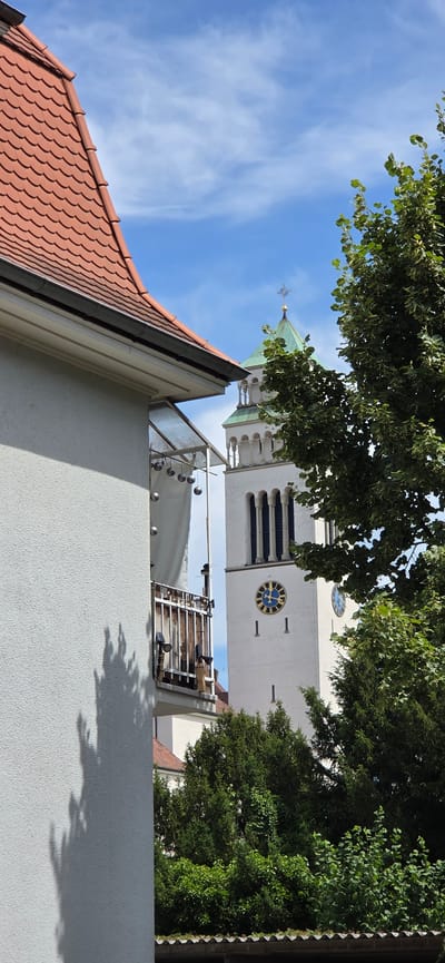 In Kehl, Germany, a white building with a textured facade and a red tiled roof is in the foreground, partially obscuring a view of a white church tower. The church tower features a green pointed roof topped with a golden cross and a weather vane. It has multiple arched windows and two visible clock faces with blue and gold details, indicating the time. A balcony with metal railings, wooden slats, and hanging decorative balls is visible on the side of the foreground building. Lush green trees, some casting shadows on the building, surround the scene, with a bright blue sky with wispy clouds above. The overall scene depicts an urban setting with architectural elements and natural foliage under a clear sky.