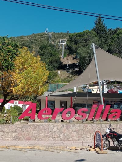 The image captures the entrance area of a chairlift attraction, likely the "Aerosilla" in Villa Carlos Paz, Argentina, given the prominent signage and the provided location context. In the foreground, large, stylized red letters spell out "AEROSILLA." Behind these letters is a building with a light-colored, sail-like canopy, which appears to be the ticket office or a related facility. Signs visible on the building include "BOLETERIA" (Ticket Office) and "PRECIOS" (Prices). To the right of the sign, a motorcycle is parked, with a nearby circular sign indicating "EXCLUSIVO MOTOS" (Exclusive Motorcycles).
The background features a steep, tree-covered hillside dotted with the towers and chairs of a functional chairlift system, ascending towards the top. The foliage on the hill is predominantly green, while some trees in the midground display vibrant yellow leaves, suggesting an autumn season. The sky is a clear, bright blue, indicating sunny weather. Overhead power lines are visible in the upper left corner. The scene is brightly lit by natural sunlight, suggesting it is daytime. The overall impression is that of a popular outdoor recreational and tourist site.