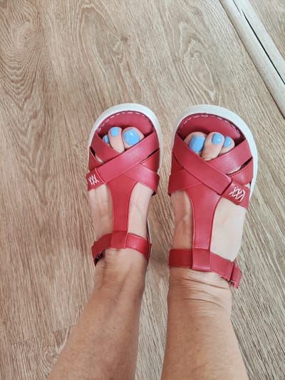 This image shows a pair of red sandals with white soles, worn by a person with light skin and blue-painted toenails. The sandals have straps that crisscross over the toes and a strap around the ankle. The person is standing on a light brown wooden floor. The scene appears to be indoors, with a reflection of what looks like a mirror or window visible in the background on the right side of the image. There is no indication that this image was taken in Gvardeysk, Russia, or any specific city.