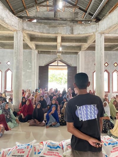 The image depicts a large gathering of people, primarily women, inside a communal building in Meunasah Ara, Indonesia, likely a Meunasah. Most women are seated on the floor, attentive, and wearing headscarves (hijabs) in various colors.
A man, seen from behind, wearing a black t-shirt with a purple geometric design, stands prominently in the center-right, holding a small dark object. To his right, several large white sacks are stacked, which are central to the activity. These bags are labeled with text including "TANGSE," "KUALITAS TERJAMIN," "BRUTTO: 5 Kg," "IDM001211076," "Berus Tangse Alen," and "ACEH RICE SPECIAL," along with "AYI & RIZKI." This indicates they are 5kg bags of rice from Tangse, presumably being distributed as aid or provisions.
The building itself features exposed concrete pillars and beams supporting a high roof of corrugated metal over wooden rafters. Natural light illuminates the interior through a large doorway at the far end and smaller, geometrically shaped windows on the upper walls, indicating it is daytime. A ceiling fan is also visible. The scene suggests an organized community event, likely a distribution of rice, taking place in a functional, unadorned communal hall. The people appear to be waiting patiently and respectfully.