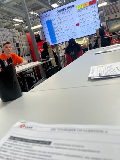 The image shows an indoor scene, likely a factory or warehouse setting, with a focus on a digital display screen and a person. The screen displays a table with columns for "Oddelen" (Department), "Problém" (Problem), "Kdo se musí zapojit do vyhodnocení problému?" (Who must be involved in evaluating the problem?), "Kdo má autorizovat implementaci navrhovaných kroků?" (Who has authorization to implement the proposed steps?), and different escalation levels (Stupeň eskalace I, Stupeň eskalace II). The text indicates an "Eskalační proces" (Escalation process). Below the screen, a person in an orange t-shirt is visible, looking towards the camera. The person has a tattoo on their arm. The background shows racks of boxes, some with the "LEAR" logo, and industrial shelving. There are also chairs, including a prominent red and black gaming-style chair. The lighting suggests it is daytime, but the specific time or weather is not discernible. The overall impression is of a work environment where processes and problem-solving are being managed. The presence of the "LEAR" logo suggests the company LEAR Corporation.