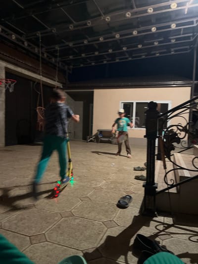 This image depicts a nighttime scene on a paved outdoor area, likely a courtyard or patio, with a building in the background. Two young boys are present. One boy is on the left, blurred due to motion, riding a yellow scooter with illuminated wheels. He is wearing a dark, patterned t-shirt and teal pants. The other boy, on the right, is also blurred and appears to be playing with a racket and possibly a shuttlecock. He is wearing a light green t-shirt, brown pants, and a white baseball cap. The environment includes a dark ceiling structure with lighting fixtures above, a basketball hoop mounted on a wall to the left, and a set of stairs with an ornate black metal railing on the right. Several pairs of sandals are scattered on the ground. The overall impression is a casual, recreational activity taking place outdoors after dark. The location is identified as Kaspiysk, Russia.