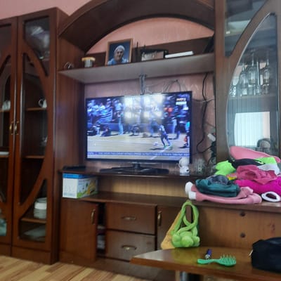 The image depicts an interior room within a residence in Kurganinsk, Russia. A large, dark brown wooden entertainment unit, composed of multiple sections including cabinets, shelves, and drawers, occupies the majority of the background.
Central to the unit is a flat-screen television displaying a soccer match in progress, featuring multiple athletes on a green field. Russian text visible at the bottom of the screen identifies the channel as "МАТЧ!" (MATCH!) and states, "ЛУИС ГАРСИА СМЕНИЛ МАТИАСА АЛМЕЙДУ НА ПОСТУ ГЛАВНОГО ТРЕНЕРА 'СЕВИЛЬИ'" (Luis Garcia replaced Matias Almeida as head coach of 'Sevilla'). A white figurine is positioned on the shelf to the right of the television.
To the left of the television, a light blue and white box labeled "D Well" rests on a lower shelf. Below it, a dark wooden cabinet door is slightly ajar, revealing internal contents. The upper left section of the entertainment unit comprises two glass-fronted cabinets containing white ceramic dishes and mugs. The upper right section features a similar glass-fronted cabinet displaying multiple inverted wine glasses.
On an open shelf above the television, a framed photograph of an older male with grey hair is visible. Adjacent to it, a small clear jar with a yellow lid, eyeglasses, and a white notebook are present.
To the right of the television, on a surface of the entertainment unit, a disorganized pile of multicolored clothing, predominantly in shades of pink, green, black, and white, is stacked.
In the lower right foreground, a portion of a dark wooden table with a metal base is visible. On its surface, a bright green plastic hairbrush and a small, cylindrical blue and silver object (possibly a lighter) are placed. A green plush toy, appearing to be a frog or turtle, with a strap, rests on the table, leaning against the entertainment unit.
The room's walls are a light salmon/pink color, possibly with a subtle texture. The floor is covered with light brown wooden laminate planks. No live individuals are present in the immediate scene, aside from those depicted on the television screen.