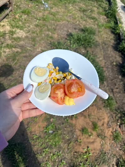 A person is holding a white plate containing sliced tomatoes, boiled eggs, corn, imitation crab meat, and a dollop of mayonnaise. A spoon with a white handle and floral design rests on the plate. The person has manicured nails with white polish. The background is a grassy area with patches of dirt, suggesting an outdoor setting. The location is Черёмушки, Russia.