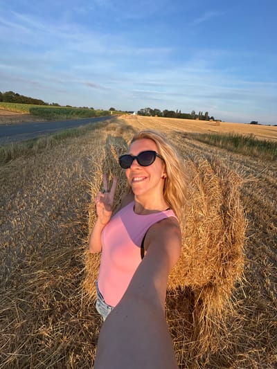 In a field of harvested crops, likely near Trouville-Alliquerville, France, a woman is taking a selfie. She has blonde hair and is wearing sunglasses and a pink tank top with denim shorts. She is smiling and holding up two fingers in a peace sign. Behind her, hay bales are stacked. The setting sun casts long shadows across the field, and the sky is a clear blue with a few wispy clouds. A paved road runs along the edge of the field in the background, with trees and more hay bales visible in the distance.