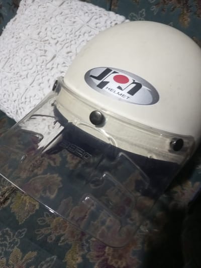 In an indoor setting within B. Nam Ngao, Laos, a white "JON HELMET" branded motorcycle helmet is depicted. The helmet features a clear plastic visor attached by three black circular fasteners, two of which are visible. The number "3820" is embossed on the visor's upper left section. The helmet rests on a dark patterned fabric, possibly upholstery, displaying teal, green, brown, gold, and purple floral or leaf motifs. Behind the helmet, a white, intricately textured or crocheted pillow or cushion is visible. No individuals are present in the scene.