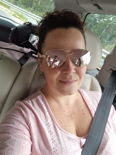 A woman with dark hair pulled up and wearing sunglasses is seen in the front passenger seat of a vehicle. She is wearing a pink, lacy top and a seatbelt is fastened across her chest. The background shows the interior of the car with bags in the back seat and greenery visible through the windows. There are reflections in her sunglasses, showing another person holding a phone. The location is not Shipka, Bulgaria.