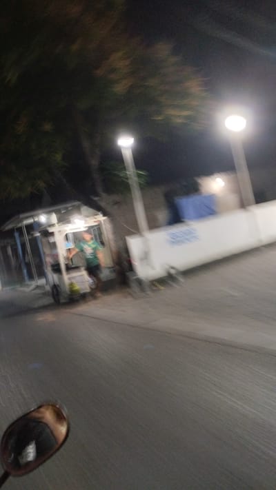 The image depicts a nighttime scene on a street in Bangsri, Indonesia. The focal point is a food cart illuminated by overhead lights. A person, likely a vendor, is standing behind the cart, wearing a green t-shirt. The cart itself appears to be made of metal and has various items displayed on it, though they are not clearly discernible due to the image's blurriness.
In the background, two streetlights are visible, casting a glow on the surroundings. A white barrier with blue lettering runs along the right side of the image, and beyond that, some buildings and trees can be seen, their details softened by the darkness. The road surface is visible in the foreground, with a motorcycle's rearview mirror reflecting a portion of the scene. The overall impression is one of a quiet, possibly late-night street scene in a local Indonesian setting. The motion blur suggests the photo was taken while moving.