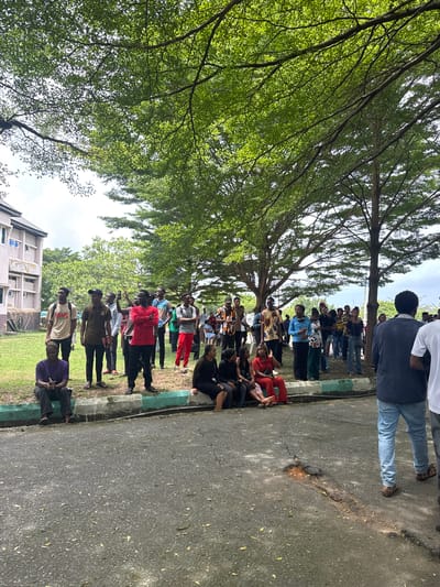 A large group of people is congregated outdoors in Eziobo, Nigeria. They appear to be mostly young adults, and they are dressed in a variety of casual clothing. Some are standing, some are sitting, and some are interacting with one another. A large tree provides shade for part of the group. The setting appears to be a public space, possibly a park or campus, with a building visible in the background. The ground is paved in some areas and covered in grass in others.