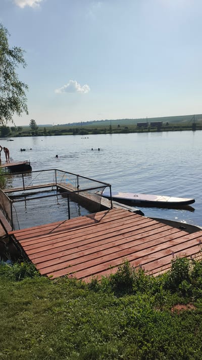 The image captures a serene summer day at a lake located in the village of Kiriushkino, Russia. The clear blue sky, dotted with a few wispy clouds, stretches over the calm, reflective water. In the foreground, a wooden dock with a red-brown finish extends into the lake, its planks laid out horizontally. Adjacent to the dock, a netted enclosure partially submerged in the water suggests a designated swimming or training area.

Several individuals are visible in the water, engaged in recreational activities. Two people are standing on a smaller, dark-colored dock to the left of the frame, while others are swimming and wading further out in the lake. A paddleboard, labeled "ALOHA," rests on the water near the main dock, indicating the possibility of water sports.

The background reveals a picturesque landscape of rolling green hills and fields, characteristic of the Russian countryside. A line of trees separates the water from the distant terrain, and faint structures, possibly buildings or agricultural outbuildings, can be seen on the horizon. The overall scene conveys a sense of tranquility and summer leisure, characteristic of a rural getaway in Kiriushkino.