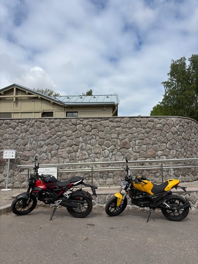 The image shows two motorcycles parked next to a stone building with a metal railing in Vyborg, Russia. The motorcycle on the left is red and black, while the one on the right is yellow and black. A sign in Russian is visible near the red motorcycle. The building appears to be made of large, irregularly shaped stones. A small structure with a metal roof is connected to the stone building. The sky is cloudy. The location is near the entrance to the "Monrepo" museum-reserve.