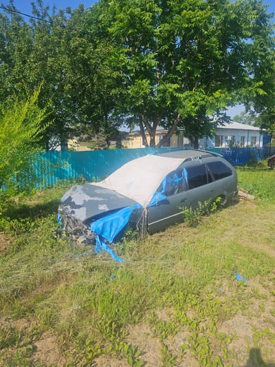In Georgievka, Russia, a partially dismantled and covered dark green station wagon sits in a grassy, overgrown area. The front of the vehicle is exposed, showing significant damage and exposed engine components. Blue tarp material is visible covering parts of the front and sides, along with a larger light-colored tarp draped over the hood and windshield. The car is positioned near a bright turquoise metal fence, with residential buildings and trees visible in the background under a clear blue sky. The overall scene suggests the vehicle is abandoned or undergoing significant repair/disassembly outdoors.