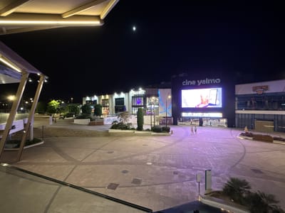 This is a nighttime, wide-angle shot of an outdoor plaza in Alcorcón, Spain. The plaza is paved with stone tiles and features several modern buildings with illuminated signage. On the left, part of an outdoor canopy with integrated lighting is visible.
Dominating the background is a large building with "cine yelmo" prominently displayed. Below this, a large digital screen shows an advertisement. To the right of the cinema, a building with "BOWLING" signage is also visible. Further to the left, other businesses like "TACO BELL" and "KFC" are indicated by their respective signs.
The scene is lit by artificial lights from the buildings and street lamps, creating a vibrant, albeit somewhat dark, atmosphere. The time of day is clearly night, with a visible moon in the dark sky. There are a few scattered individuals walking across the plaza, suggesting a moderate level of activity. The overall mood appears calm and urban, typical of a commercial or entertainment district at night. The smooth paving, modern architecture, and the presence of entertainment venues like a cinema and bowling alley point to a leisure or shopping complex.