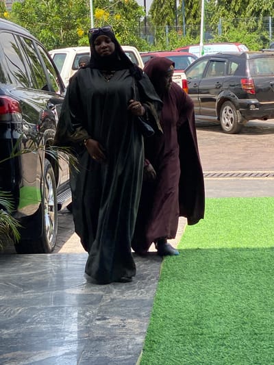 Two individuals, dressed in traditional Islamic attire, are captured in a medium shot. The person in the foreground, wearing a dark green abaya and hijab, is facing the camera with sunglasses perched on her head. She is also wearing a ring on her left hand. Behind her, another person, also in Islamic attire, is partially visible. The scene appears to be an outdoor setting, with a paved walkway in the foreground, artificial grass to the right, and parked vehicles in the background. The lighting suggests it is daytime, with dappled sunlight casting shadows. The overall mood is neutral. There are no visible text elements in the image.