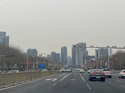A wide city road with multiple lanes is filled with cars moving towards a cluster of modern high-rise buildings in the distance. The sky is overcast, creating a muted light over the urban landscape. Bare trees line the sides of the road, and traffic lights hang over the lanes. A median barrier separates the opposing directions of traffic. A sign in Chinese can be seen next to the leftmost set of traffic lights.