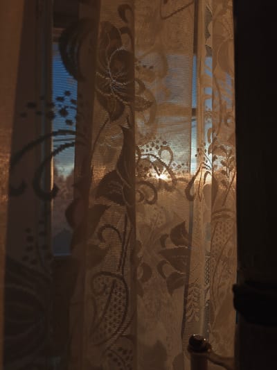 The image depicts a window with lace curtains, illuminated by the warm glow of the setting or rising sun. The sun's rays are filtering through the delicate fabric, casting intricate floral patterns and shadows onto the curtains and the interior of the room. The sky visible through the window appears to be a muted blue, suggesting either early morning or late evening. The overall mood is serene and tranquil. The location in the city of Hadiach, Ukraine is not explicitly discernible from the visual elements within the photograph. There are no people or active interactions visible in the scene. The environment appears to be an indoor domestic setting, with the focus on the interplay of light and shadow on the window and curtains.