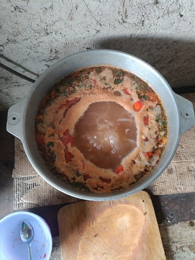 The image shows a large metal pot filled with a simmering stew. The stew is a reddish-orange color and appears to contain chopped tomatoes and herbs. Bubbles rise to the surface, indicating it is actively cooking. The pot has two handles and is placed on what appears to be a rustic wooden surface, possibly a table or counter.
In the lower left of the frame, a light purple bowl with a spoon inside is visible. The bowl shows some faint red stains, suggesting it was used for serving the stew previously. To the right of the pot, a wooden cutting board rests on the same surface.
The background consists of a textured, light-colored wall that looks somewhat rough and aged. The overall impression is of a simple, home-style cooking environment. The lighting suggests it is daytime, but no specific time can be determined. The setting, with the rustic surfaces and textured wall, could be interpreted as a kitchen or outdoor cooking area in a rural location. No people are visible in the image. There is no visible text. The location context is Tash-Dobo, Kyrgyzstan, which aligns with the rustic and possibly traditional elements of the scene.