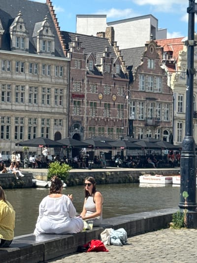 In this media file, people are enjoying a sunny day by a canal in Ghent, Belgium. The scene depicts a charming canal-side with historic brick buildings adorned with numerous windows. Several individuals are seated on a stone ledge by the water, with some appearing relaxed and others engaged in conversation.
One woman, wearing a striped top and sunglasses, is seated, while another, in a white long-sleeved shirt, is in the foreground, facing away from the camera. A person in a yellow top with a polka dot pattern is also visible. Clothing items, including a blue shirt and a red jacket, are casually placed on the ledge.
The activity appears to be a leisurely outing, possibly a break from sightseeing or a casual get-together. The presence of umbrellas over an outdoor seating area suggests a cafe or restaurant. The lighting indicates it is daytime, likely with pleasant weather given the outdoor activities and attire. The architecture of the buildings, with their distinctive gabled roofs and brick facades, strongly points to a European city, consistent with the provided location context of Ghent, Belgium.
A sign with text is partially visible on a building, reading "Frederi Dewaele" and "Red." The overall mood conveyed is one of relaxation and enjoyment of a beautiful day in a historic urban setting.